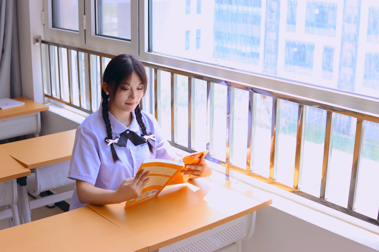 A young woman reads a book by the window in a bright, sunlit classroom.